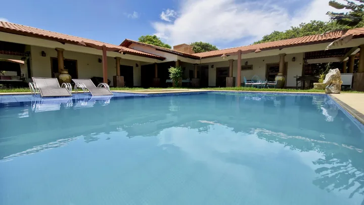 Piscina em frente à casa e varanda, com espreguiçadeiras ao fundo.