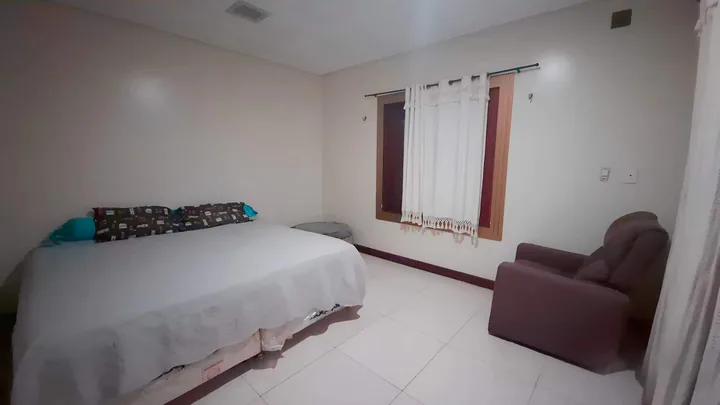 Quarto 4 com cama de casal, poltrona marrom e janela com cortina.